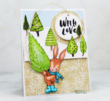 Card with the bunny colored in orange with a blue scarf and boots standing among green watercolor pine trees, a kraft circle stamped 'with love' hanging above on twine.