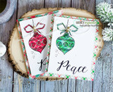 Two plaid cards stacked, one with a red ornament and one with a green ornament, both tied with twine, on a wood background.