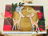 Card with red buffalo plaid and a wood grain circle, two deer coloured in tan with green foliage sprigs and a white believe die cut across the front.