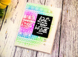 Rainbow blended panel with the knit pattern masked in white, holding a small black label with a scripture sentiment, set on a pale wood background.