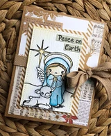The angel and lamb on a kraft card layered with music-sheet paper and a bow, reading 'Peace on Earth.'