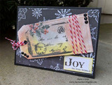 Navy snowflake card with the aged tag, a holly sprig, red and white bakers twine, black gems, and a Joy to the World label.