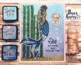 Card with tall stamped seaweed, a jellyfish and the mermaid in blue and gold, beside distress ink pads.