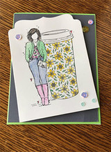 Card on a wood desk with a fold-back flap, a girl in a green jacket and pink boots leaning on a tall floral travel cup, enamel dots along the edge.