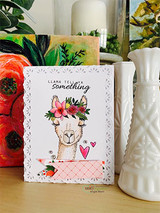 The floral-crown llama on a clean white card with a pink diamond panel — 'Llama tell ya something.'