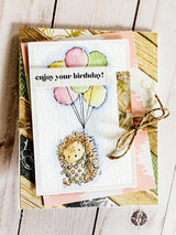 Layered card on gold and pink papers: the hedgehog with pastel balloons above “enjoy your birthday!” tied with twine.