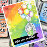 Rainbow watercolor panel with the hedgehog holding uncolored balloons above a black “enjoy your birthday!” banner, styled with ink pads.
