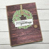 Dark wood grain card with a green holly wreath hung from gold ribbon and "Season's Greetings" stamped on a white strip.