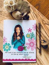 Floral card with the girl in a teal top holding a cross and 'The Lord is my strength and my shield.'