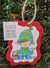 An ornament shaped tag hung on a tree, stamped with the elf boy colored in blue and green, beside a small panel reading 'DO NOT OPEN UNTIL CHRISTMAS'.