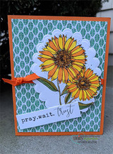 An orange card on a green leaf print with the blanket flowers colored in golden yellow on a scalloped cloud panel, tied with orange ribbon, above the sentiment 'pray. wait. trust'.