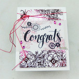 Card with 'Congrats' stamped in navy across a white panel over a background of black and pink outlined flowers, wrapped with pink thread and finished with pink gems.