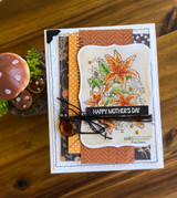 A rust and black layered card with orange lilies colored on a scalloped panel, tied with black twine.