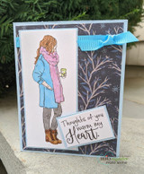 Handmade card featuring the autumn girl bundled in a coat and scarf holding a warm drink, in a cool blue winter scene, stamped with 'Thoughts of you warm my heart'.