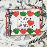 Card bordered with stamped red and green apples around "Teach Love Inspire", tied with a gingham bow on a red base.