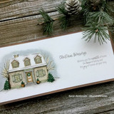 A cozy home framed by snowy evergreens hand-stamped on a card and lettered "Wishing you the warmth of home."