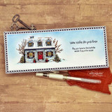 A cozy home framed by snowy evergreens hand-stamped on a card and lettered "Wishing you the warmth of home."