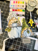 Watercolored gnome pair with pumpkins fussy-cut on a craft desk with paints