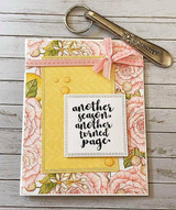 Card with a frame of peach roses and lemons around a gold embossed center panel reading 'another season, another turned page', finished with a pink ribbon.