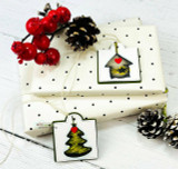 White polka-dot gift box with a house-ornament tag and a small tree ornament, beside pinecones and berries.