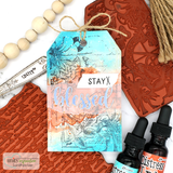 Mixed media sample tag in teal and coral distress ink with the gladiolus stamped into the background, styled with ink bottles and rubber texture plates.