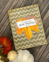Orange maple leaf on a tan chevron card with sequins and 'In every thing give thanks', styled with pumpkins.