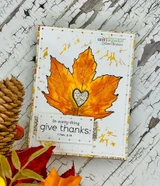 Golden orange maple leaf with a brown glitter heart on a gold-splattered white card, stamped 'In every thing give thanks'.