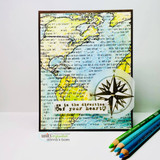 Card on white with colored pencils beside it, the inky world map background stamped over green and blue watercolor, with a compass rose die cut and a sentiment strip at the bottom.