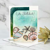 Card with shells coloured in pale pink and blue sitting on a sandy beach with a teal sky, sentiment "Let's Shellebrate!"