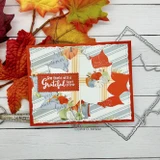 Sample card on white wood surrounded by red and orange maple leaves. Fall leaves and pumpkins layer over a soft striped panel, finished with a small gratitude tag and stitched dies.