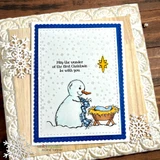 Card in a wood and snowflake frame, a navy scalloped panel with the snowman and manger stamped in soft blue, a gold star, and "May the wonder of the first Christmas be with you."