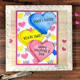 Card with three hearts coloured blue, yellow and pink on an xoxo-printed panel in a pink frame, sentiments "HUGS & KISSES," "YOU'RE SWEET" and "HAPPY VALENTINE'S DAY."