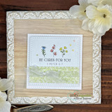 Wood-framed card with a green floral panel, sentiment 'HE CARES FOR YOU 1 PETER 5:7' above small yellow and pink wildflowers.