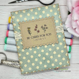 Sage polka-dot notebook cover with a kraft label stamped 'HE CARES FOR YOU 1 PETER 5:7' beside small pink flowers.