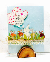 Blue card with a heart of red hearts above bunnies in a grassy border and 'oh Hello Spring.'
