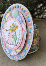 Oval easel card with the bunny and daisy over pastel eggs and pom-pom trim.