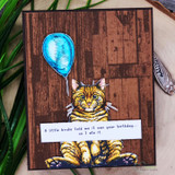 Dark brown card with the wood planking stamped in deep walnut, a tabby cat holding a blue balloon on a white panel and a cheeky birthday greeting below.