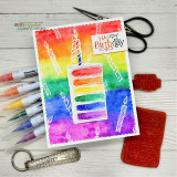 Rainbow watercolor card with a white panel: the cake slice in rainbow layers beside “HAPPY BIRTHDAY,” red rubber stamps and markers alongside.