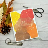Handmade sample card using the Abstract Splotch masking stencil — overlapping organic paint-blob shapes blended in warm autumn yellows, oranges and browns, styled with fall leaves and a pinecone.
