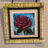 Framed art piece of a red rose on blue over a yellow crackle mat.