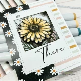 Card with a colored daisy in a frame and 'There' over a striped background with fresh daisies.