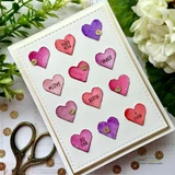 White stitched card with a scatter of purple and magenta hearts each stamped with a word such as "HUGS" and "BESTIE", styled with vintage scissors and white blooms.