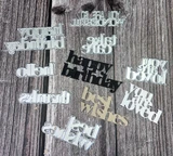 The Happy Hello metal word dies from Unity Stamp Co laid on weathered wood — die-cut sentiment words including 'happy birthday', 'hello', 'thanks', 'best wishes', 'take care' and 'you are loved' in silver.