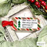 Striped holiday tag with a stamped tree and “The tree isn't the only thing getting lit this year,” tucked into a whitewashed frame with greenery.