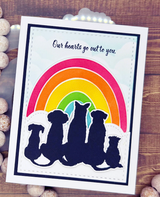 Handmade card with a bold layered rainbow in red, orange, yellow, green and blue arching over navy silhouettes of dogs and cats sitting in a row, the sentiment 'Our hearts go out to you.' on a white panel over a wood background.