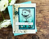 Sample card with the bear cub stamped in black line art on a teal panel with white heart accents, layered over vintage collage paper and a turquoise envelope.