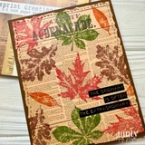 Newsprint card layered on brown, stamped with distressed leaves in red, green, rust and brown, with black label strips laid across it.
