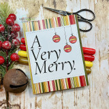 Card with 'A Very Merry.' in black on a stitched white panel with three hanging red and gold ornaments, over red, green and gold striped paper.
