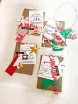 A cluster of kraft and white gift tags on twine, including the Reindeer Express label with naughty and nice checkboxes and the nativity tag, finished with a red die cut word.