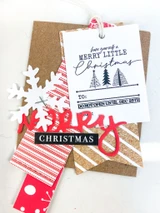 A tag stamped with the Merry Little Christmas label and its trio of trees, layered with a white snowflake, a red die cut word and striped paper.